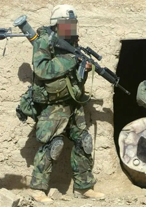 A soldier in camouflage gear stands with a rifle in front of a rugged wall, appearing alert and ready for action.