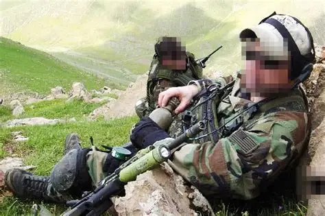 Two military personnel resting on rocky terrain, camouflaged in military gear, with one soldier holding a rifle fitted with a green tactical light.