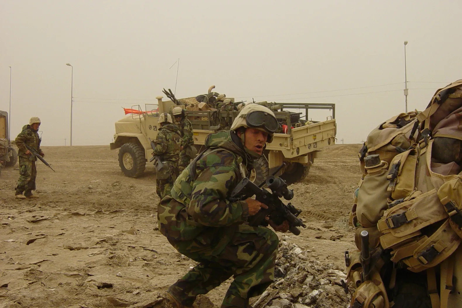 Military personnel in camouflage uniforms maneuvering in a dusty environment, with military vehicles in the background.