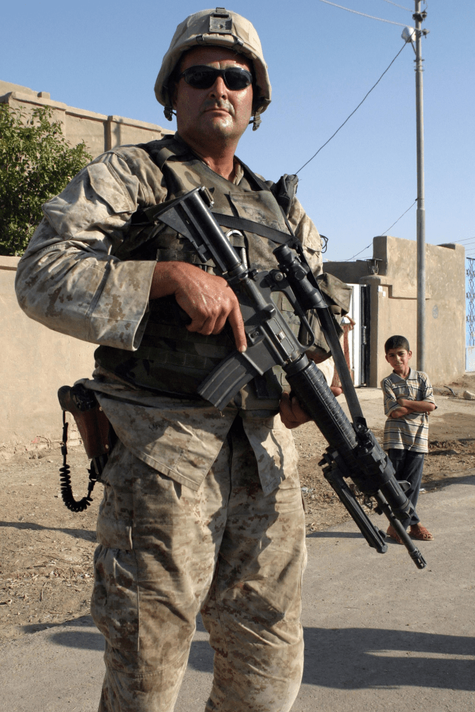 A soldier in military camouflage with sunglasses stands holding a rifle, with a building and a child in the background.