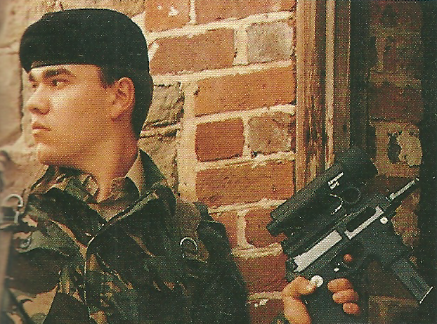 A young soldier wearing camouflage attire, holding a firearm with an Aimpoint sight, stands against a brick wall.