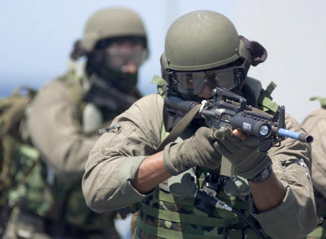 A soldier in tactical gear aims a weapon while wearing a helmet and protective goggles, with other team members in the background.