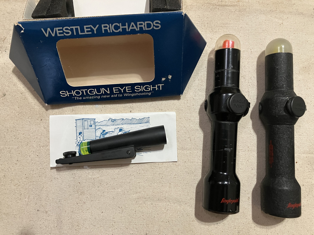 Westley Richards Shotgun Eye Sight packaging alongside two Singlepoint optics, one with a red lens and the other with a yellow lens, and an instruction booklet laid out on a beige fabric background.