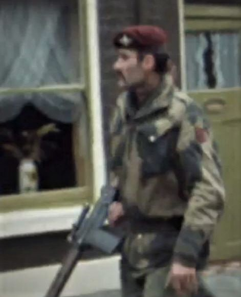 A soldier wearing a beret and camouflaged uniform, holding a firearm, walking past a house with an open window.