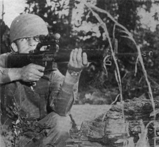 Soldier aiming a rifle equipped with an optical sight, crouching behind a log in a wooded area.