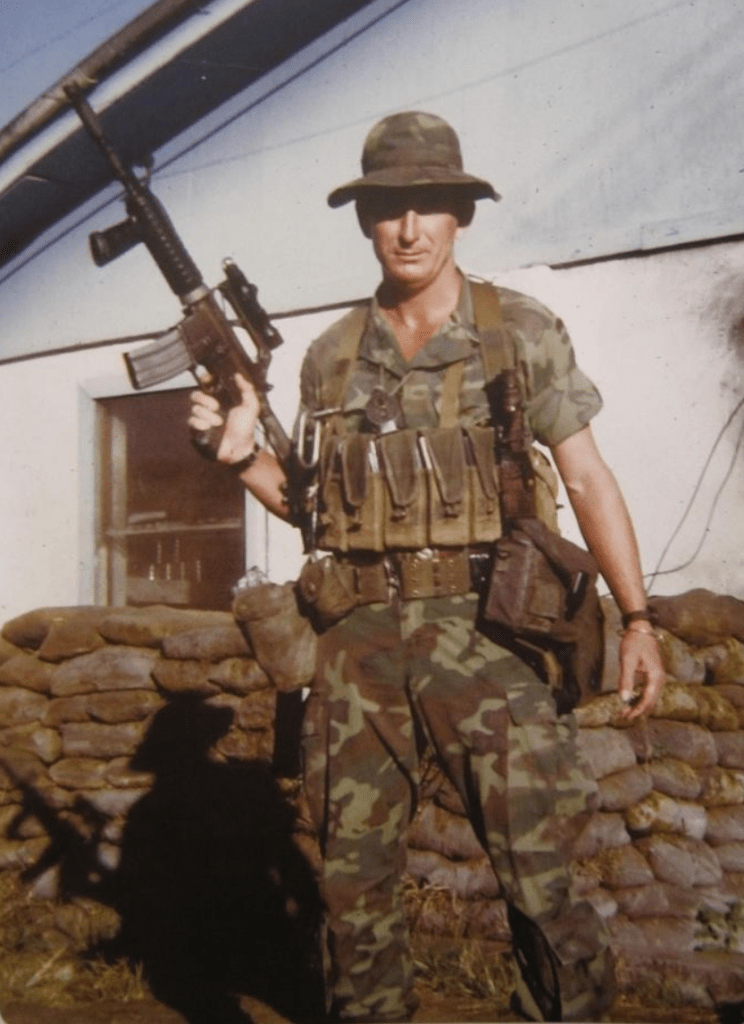 A soldier in camouflage clothing stands in front of a building, holding an assault rifle and equipped with tactical gear.