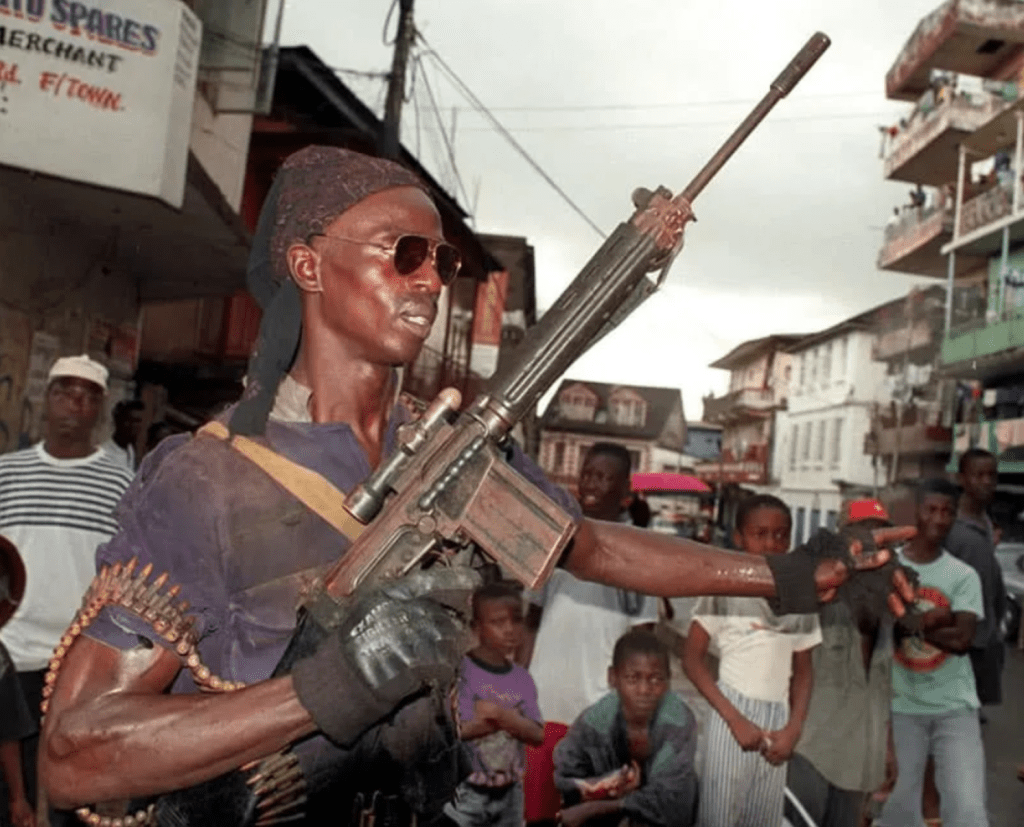 A man holding a rifle and wearing sunglasses stands in a street surrounded by onlookers, showcasing a tense atmosphere.