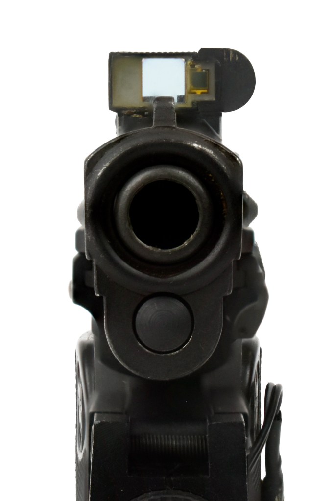 Close-up view of a firearm barrel with a red dot sight mounted on top, showing the sight's aiming window and reflection effects.
