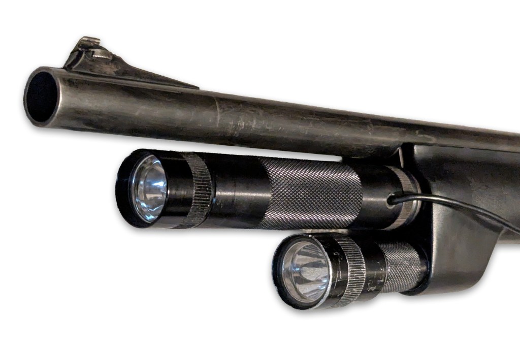 Close-up of a flashlight mounted on a shotgun barrel, showcasing two halogen bulbs with a remote pressure switch attached.