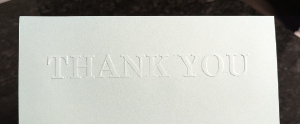 Close-up of an embossed thank you card with the words 'THANK YOU' in white lettering on a light blue background.