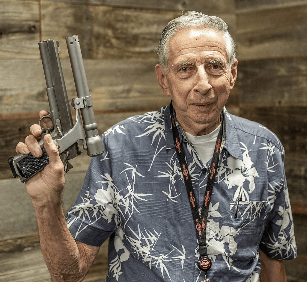 An older man in a floral shirt holding a modified handgun with a barrel attachment, standing in front of a wooden background.