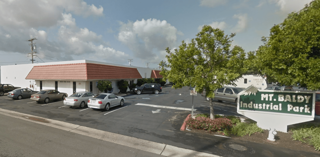 View of Mt. Baldy Industrial Park featuring buildings with red rooftops, parked cars, and greenery.